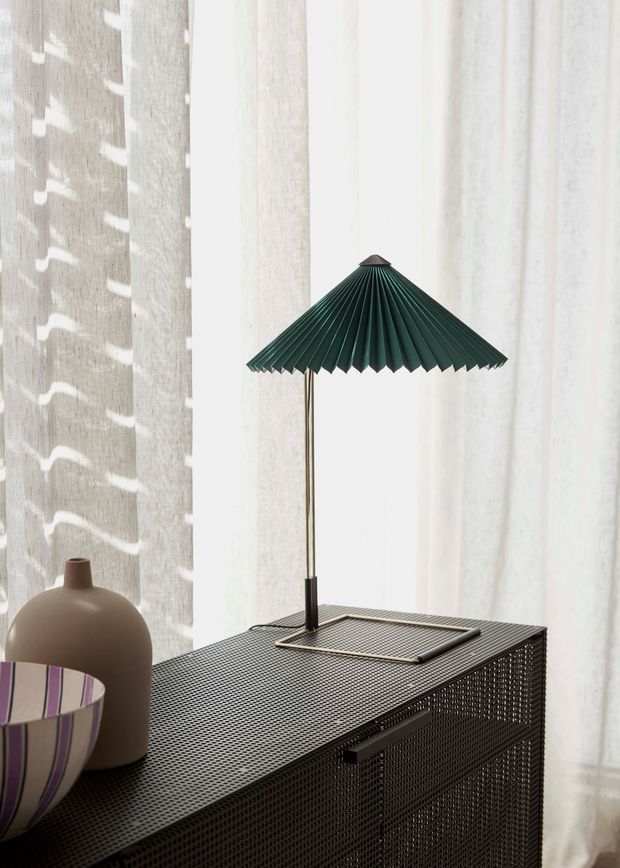 A close-up interior vignette featuring a dark woven or perforated storage cabinet with a slim green pleated table lamp on top. Sheer white curtains fill the background, while a matte taupe sculptural vase and a partially visible striped vessel add decorative accents. The composition is softly lit and refined, with strong contrast between the dark furniture and bright curtains.