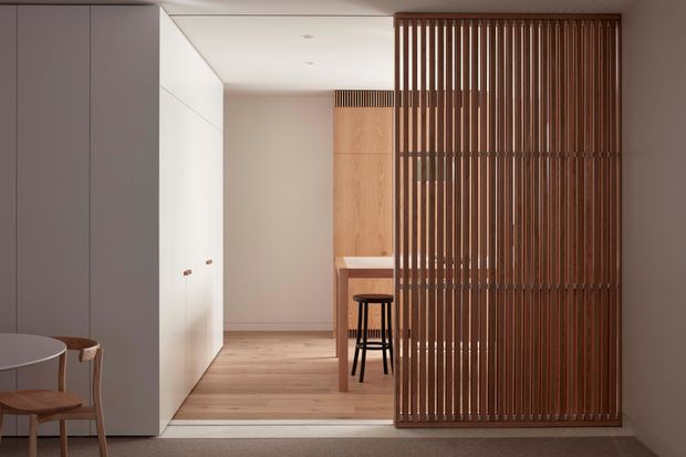 A refined contemporary interior viewed through a slatted timber room divider. The foreground contains pale built-in cabinetry and a small round table with timber chairs, while beyond the screen is a compact timber-lined workspace or kitchen-like nook with a stool. Pale timber flooring, soft blush-beige walls, white cabinetry, and warm vertical timber battens create a calm, minimalist composition.