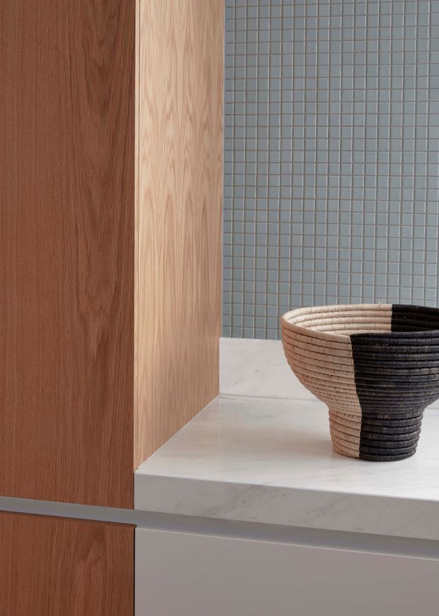 Close-up detail of a refined bathroom vanity or built-in joinery area, featuring warm vertical timber cabinetry, a pale marble or stone benchtop with a stepped edge, a muted blue-grey small-format tiled wall, and a black-and-natural woven vessel basin. The composition is minimalist and material-focused, with no broader room context visible.
