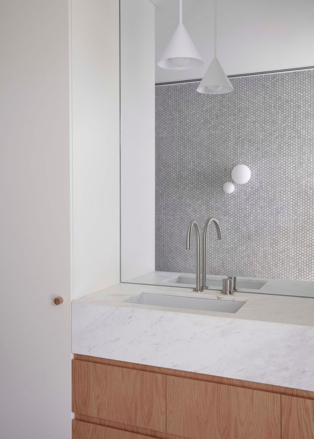 A refined minimalist kitchen wet-bar area with white flat-panel cabinetry, a pale veined stone benchtop, integrated rectangular sink, brushed metal gooseneck tap, and a full-height small hexagonal mosaic tile splashback in soft grey and white. Two sculptural white pendant lights hang above the counter, while warm timber drawer fronts add subtle contrast.