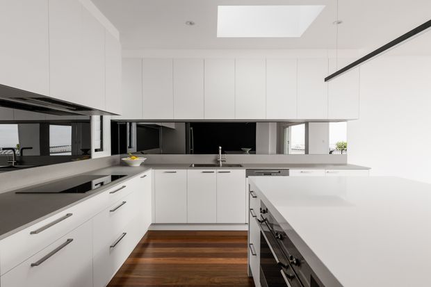 Bright contemporary white kitchen with extensive handleless and streamlined cabinetry, dark grey benchtops, a large central island with integrated cooking appliances, mirrored splashback, timber flooring, and a ceiling skylight. The kitchen opens visually toward adjacent spaces through the reflective splashback and rear opening.