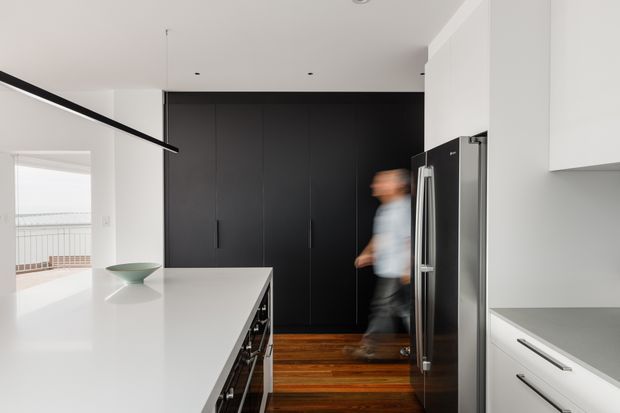 A bright, minimalist contemporary kitchen with a large white island, dark integrated cabinetry along the rear wall, white perimeter cabinetry, a stainless-steel double-door refrigerator, and warm timber flooring. A slim linear pendant light hangs above the island, with balcony glazing visible at the left.