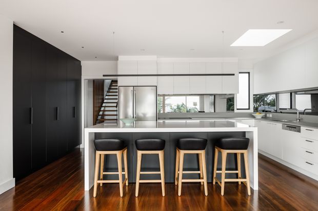 A bright, contemporary open-plan kitchen with a large white island and four bar stools. The room features extensive white cabinetry, a dark full-height storage wall, stainless-steel appliances and work surfaces, dark polished timber flooring, a glass-panelled staircase, and a large skylight. Windows and glazing bring in natural light and views of greenery.