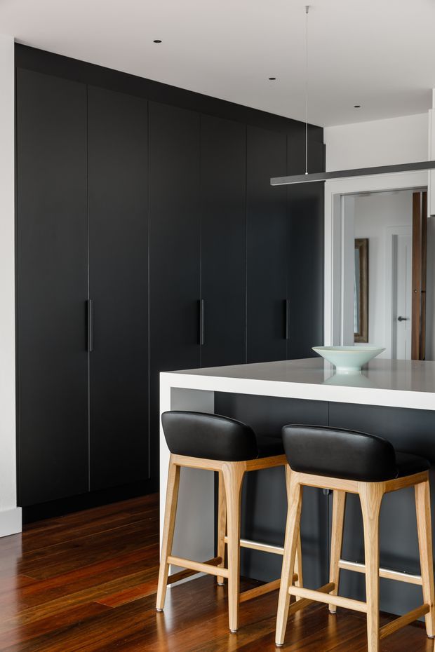 A sleek contemporary kitchen with a long wall of matte black full-height cabinetry, integrated vertical pulls, and a white island benchtop with a dark base. Two black leather and light timber bar stools sit at the island. Dark polished timber flooring, white walls and ceiling, recessed downlights, and a slim linear pendant create a high-contrast minimalist composition. A hallway and internal doors are visible beyond the kitchen.