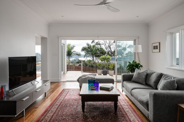 Bright contemporary living room with white walls, timber flooring, a large patterned area rug, grey sectional sofa, coffee table, television console, ceiling fan, and wide glazed doors opening to a balcony with leafy views.