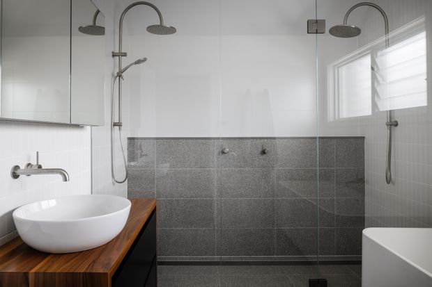 Contemporary bathroom with a large frameless glass walk-in shower, dual rainfall and handheld shower fixtures, grey tiled walls and floor, a white vessel basin on a dark timber vanity, and a partially visible freestanding white bathtub. Natural light enters through a frosted louvre window.