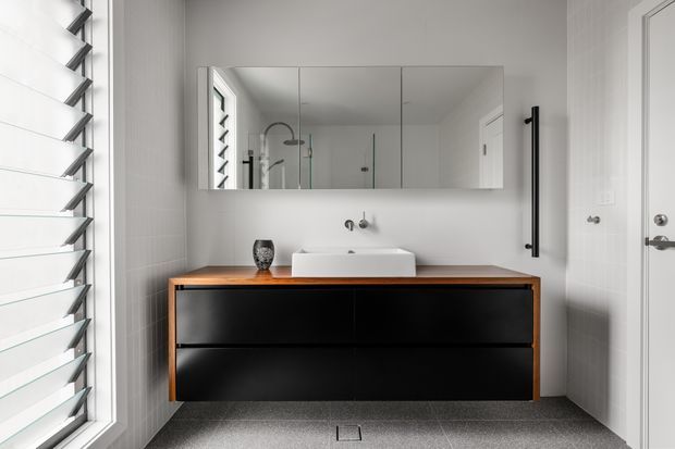 Contemporary bathroom featuring a wide floating black vanity with warm timber benchtop, white vessel basin, wall-mounted tapware, mirrored cabinetry, frameless glass shower enclosure, and a louvred window. The palette is predominantly white, grey, black, and timber, with clean-lined fittings and strong natural light.