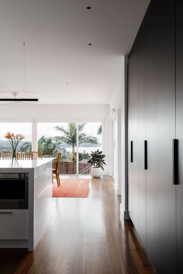 A bright, open-plan contemporary kitchen and dining area with a long white island, integrated dark oven, timber dining table and chairs, and full-height glazing opening toward a leafy coastal outlook. A large bank of tall dark cabinetry with long black handles lines the right wall. Warm timber flooring, white walls and ceiling, recessed downlights, a slim linear pendant and a red-orange rug add contrast.