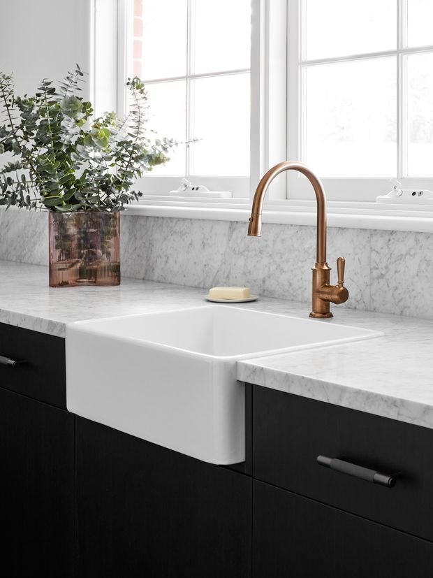 Close-up of a refined kitchen work zone featuring a deep white farmhouse-style apron-front sink, white marble benchtops and matching marble splashback, dark timber cabinetry, and a curved aged-brass mixer tap. Large white-framed windows provide abundant natural light, while a copper-toned vase with greenery adds a small decorative element.