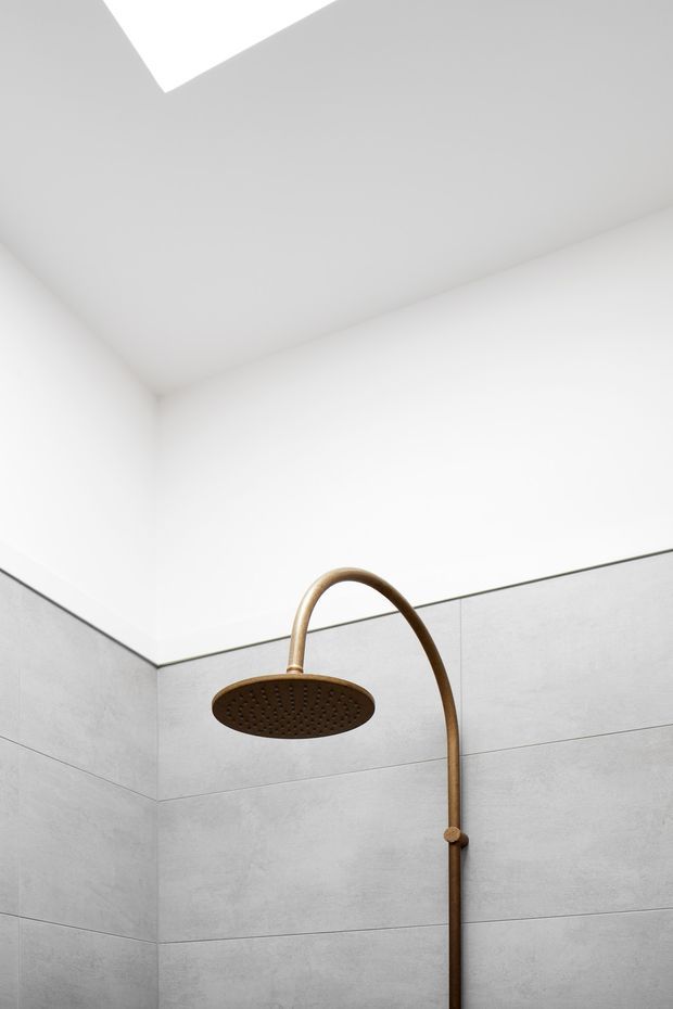 Close-up architectural photograph of a minimalist shower enclosure. Large light-grey wall tiles with horizontal grout lines surround a curved brushed bronze/gold shower arm and round rainfall showerhead. The upper walls and ceiling are white, with a bright rectangular skylight or ceiling opening visible above, creating a clean, softly lit atmosphere.