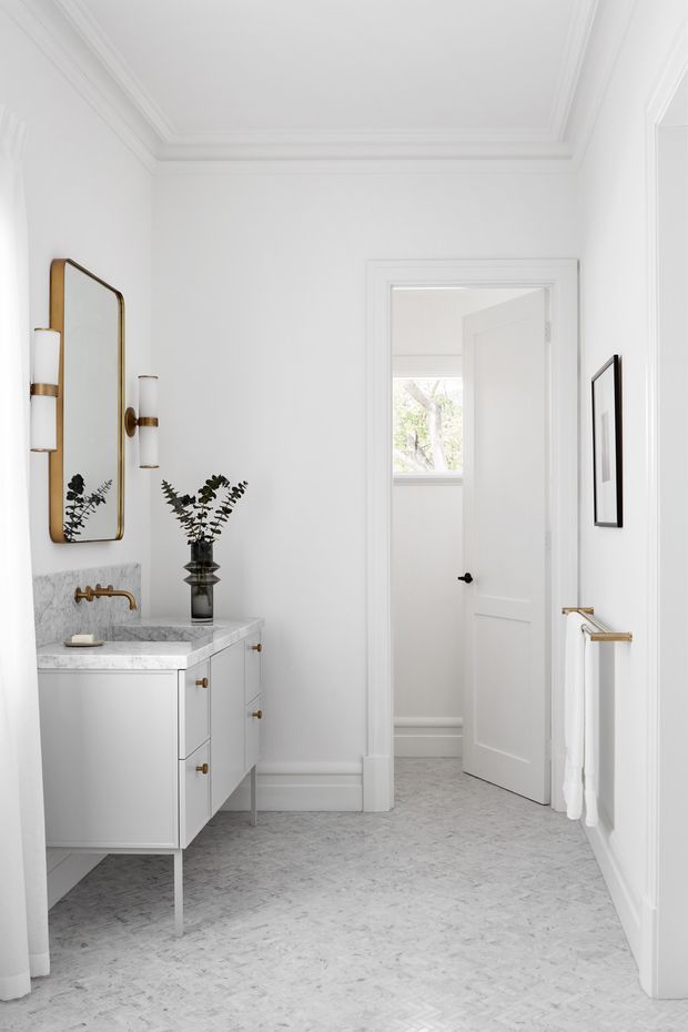 Bright, refined bathroom featuring a white floating vanity with marble countertop and integrated backsplash, brass wall-mounted faucet, framed brass mirror, wall sconces, marble-look herringbone floor, white architectural mouldings, and a doorway leading to an adjacent room. The space has a clean modern-classic aesthetic with subtle botanical styling.