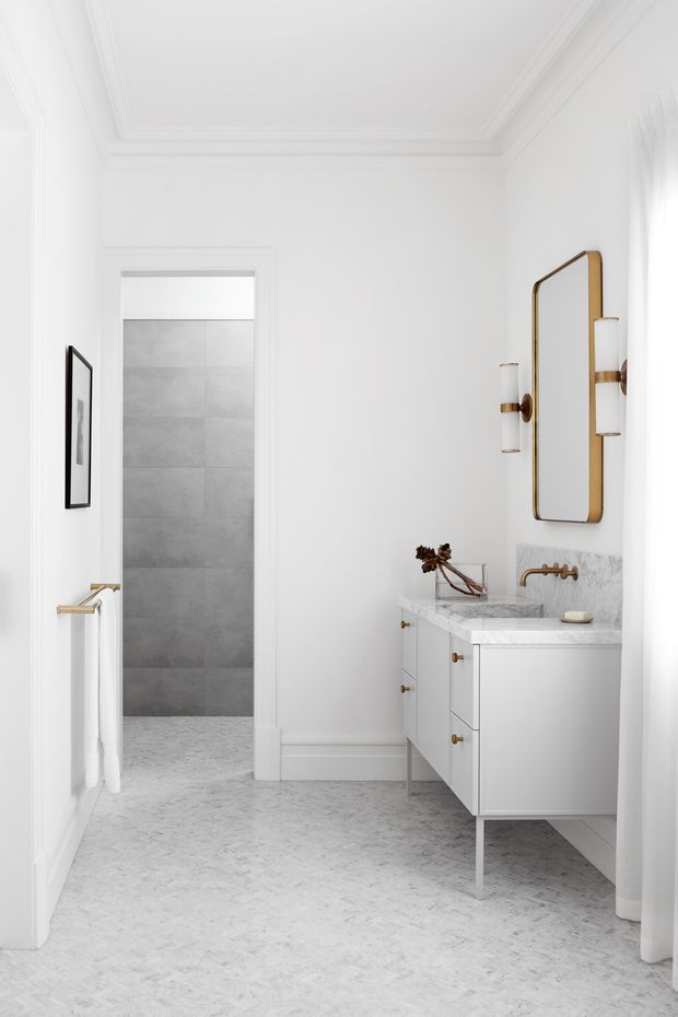 Bright, refined bathroom interior featuring a white floating vanity with pale marble-look benchtop, brass wall-mounted tapware and hardware, a large rounded-rectangle mirror, white wall panelling with decorative ceiling cornices, and a light patterned floor. A tiled shower area is visible through the doorway, with sheer curtains at the right edge and a towel rail on the left wall.