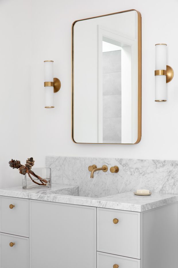 A refined bathroom vanity vignette with a white built-in cabinet, white marble countertop and integrated basin, wall-mounted brass faucet, rounded-rectangle brass-framed mirror, and matching cylindrical brass-and-white sconces. The palette is light and restrained with warm metallic accents and subtle natural decoration.