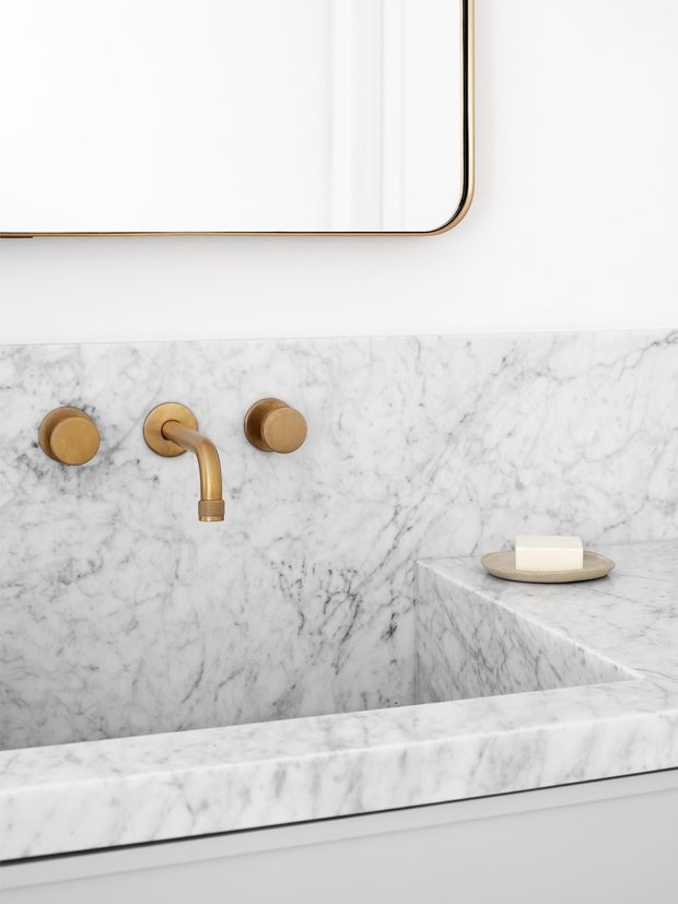 Close-up of a refined bathroom vanity featuring a wall-mounted brushed gold tap with matching circular controls, a white and grey veined marble backsplash and integrated basin, and a minimalist rounded-corner mirror with a thin gold frame. A small soap dish sits on the marble ledge.