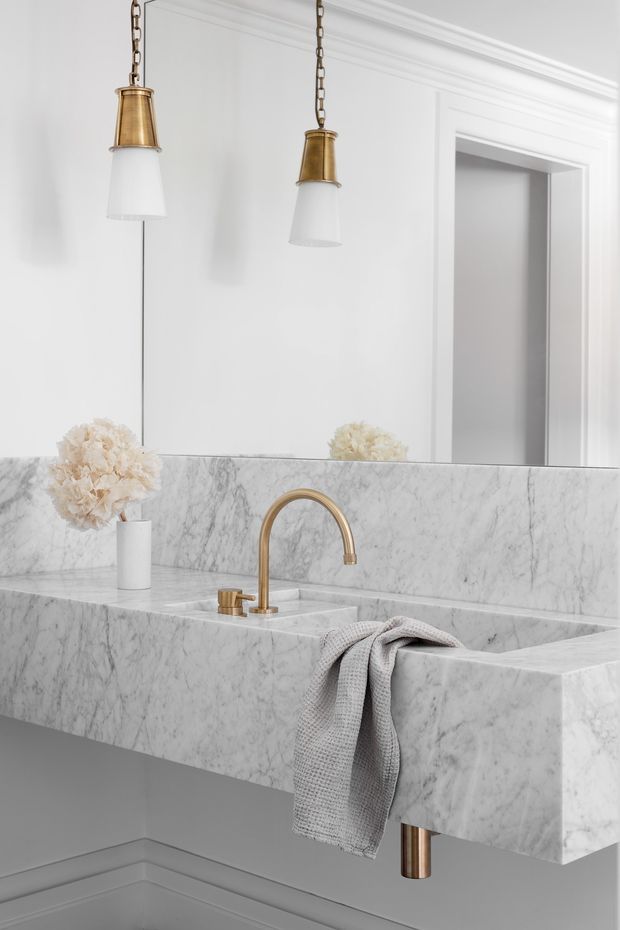 A bright, refined bathroom vanity with a large wall mirror, white painted walls and decorative trim, a thick white-veined marble vanity top and integrated basin, and brushed brass/gold tapware and pendant lights. A pale grey hand towel and small floral arrangement add soft accents.