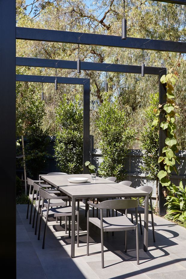 A contemporary outdoor dining terrace with a long rectangular table and multiple chairs beneath a dark steel pergola. Slim cylindrical pendant lights hang from the overhead beams, while dense planting and climbing vines create a green garden backdrop. Large grey paving slabs and strong sunlight with geometric shadows define the space.