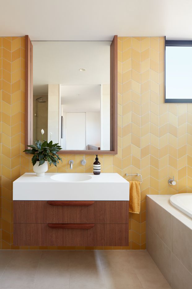 Bright contemporary bathroom featuring a floating timber vanity with a white integrated basin, large wood-framed mirror, and full-height mustard-yellow geometric wall tiles. A freestanding-style bath is partially visible at right, with a towel rail, indoor plant, and simple wall-mounted fixtures. The room has warm timber and yellow tones balanced by pale neutral floor and ceiling surfaces.