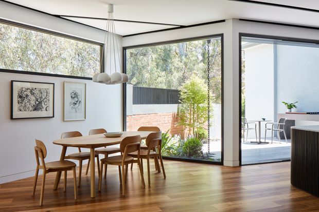 Bright contemporary dining area with a timber dining table and chairs, white walls, warm timber flooring, black-framed corner glazing, and a sculptural pendant light. The glazing opens views to a planted courtyard and an adjacent outdoor seating area, while framed artwork and minimal built-in detailing add a refined finish.