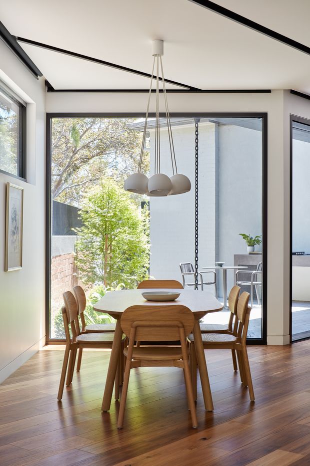 A bright contemporary dining area with a round light-coloured dining table and sculptural upholstered wooden chairs, positioned beside large black-framed glazed doors opening to a small courtyard or patio. The room has warm timber flooring, white walls and ceiling, black linear ceiling detailing, and a modern multi-arm pendant light. An outdoor seating area and planting are visible beyond the glazing.