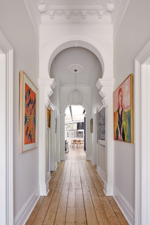 A long, elegant interior hallway with white painted walls, ornate decorative plaster ceiling detailing, elaborate archways, classical columns and mouldings, and framed colourful artwork along both sides. The hallway has wide-plank natural timber flooring and leads toward a bright dining area with modern furniture at the far end.