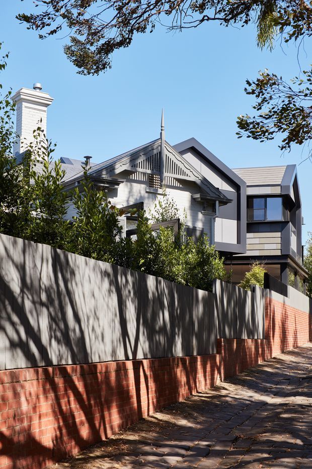 Street-level exterior view of a substantial residential home combining a traditional gabled brick-and-rendered facade with decorative timber gable detailing and a prominent chimney, alongside a newer contemporary wing with sharp angular roof forms, dark framing, large glazing, and horizontal cladding. A tall boundary wall uses red brick at the base and dark grey panels above, with a paved footpath and mature planting in the foreground.