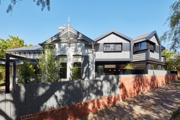 Street-facing exterior view of a contemporary multi-volume residence combining a restored or retained gabled heritage-style dwelling with a modern addition. The facade features dark cladding, large black-framed windows, projecting gabled forms, and a red-brick boundary wall with a dark vertical-slat fence. Mature landscaping and a covered entry or carport structure are visible.