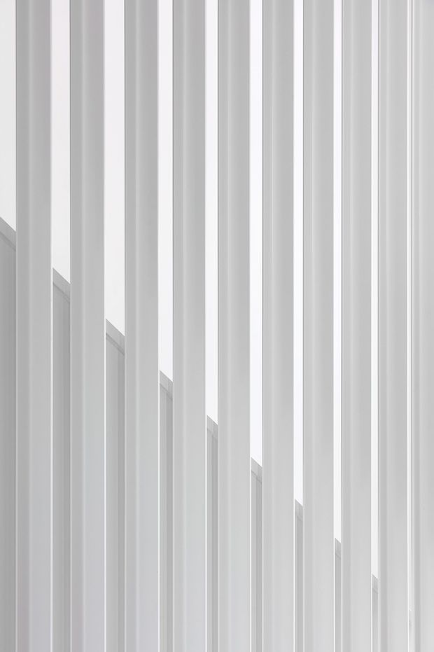 Abstract close-up architectural view of repeating tall, narrow white vertical fins or slatted elements. The elements create strong linear rhythm and layered depth, with a stepped diagonal termination receding across the composition. The image is monochromatic, bright, and highly minimalist, with no identifiable room or furnishings visible.