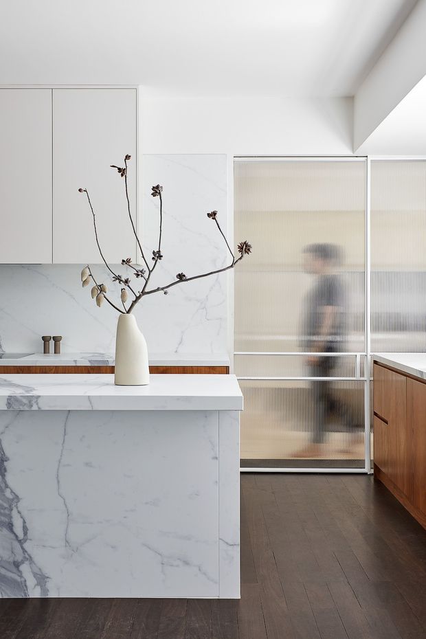 A refined contemporary kitchen with a large white marble-look island and matching full-height splashback. Minimal white cabinetry lines the back wall, while warm timber cabinetry runs along the right side. Ribbed translucent glass sliding doors separate the kitchen from an adjacent space, and dark timber flooring provides contrast. A sculptural white vase with dried branches is placed on the island.