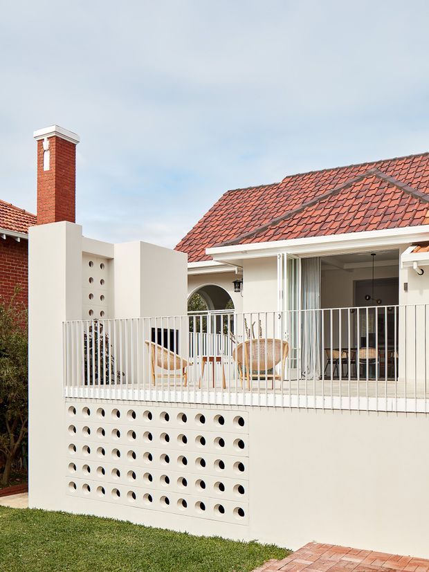 Exterior view of a renovated residential terrace or bungalow featuring a red terracotta tiled pitched roof, red brick chimney, cream rendered walls, a perforated breeze-block privacy wall, and a raised balcony or verandah with white metal balustrading. The covered outdoor area contains light timber and woven seating, with sliding doors opening into the home. The composition highlights the facade, roofline, balcony and distinctive patterned masonry screen.