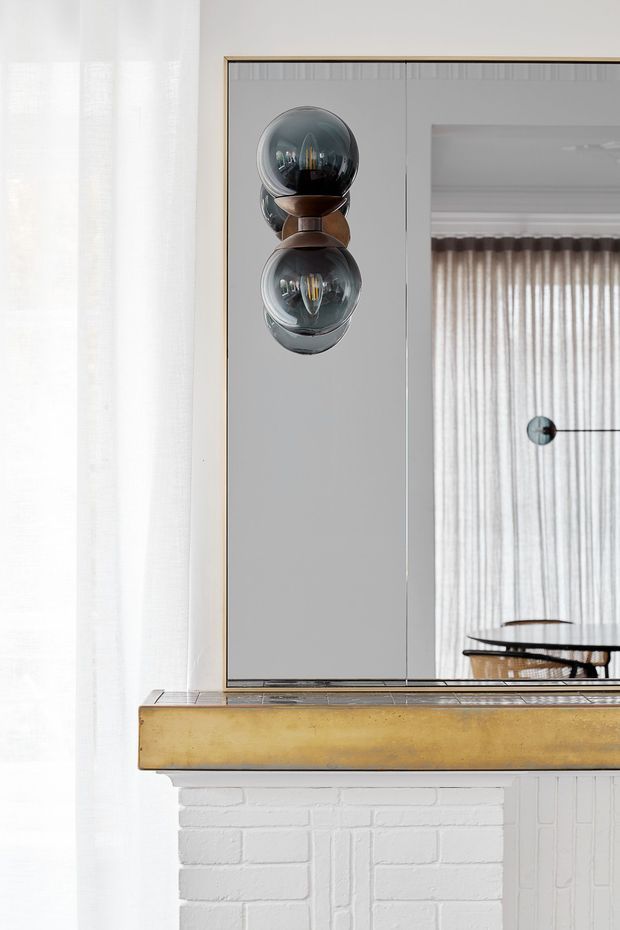 Close-up interior detail of a refined living or dining area featuring a large framed wall mirror above a white-painted brick fireplace mantel. A sculptural double-globe wall light with smoked glass and bronze-toned fittings is mounted on the mirror. The reflection shows sheer curtains, a dining table, and chairs, while a brass-toned mantel shelf adds warmth against the predominantly white and grey interior.