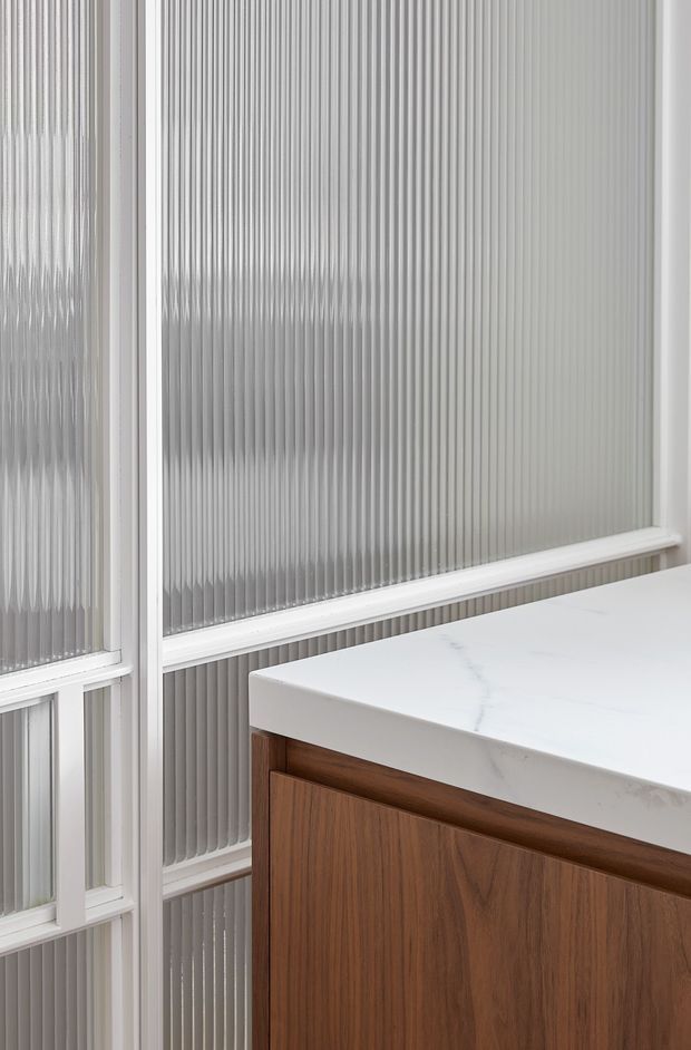 Close-up architectural detail of a white-framed window with vertically ribbed translucent glass beside a fixed wood-grain cabinet or island. The cabinet has warm medium-brown timber cabinetry and a thick white engineered-stone countertop with subtle grey veining. The image is tightly cropped, with no broader room context visible.
