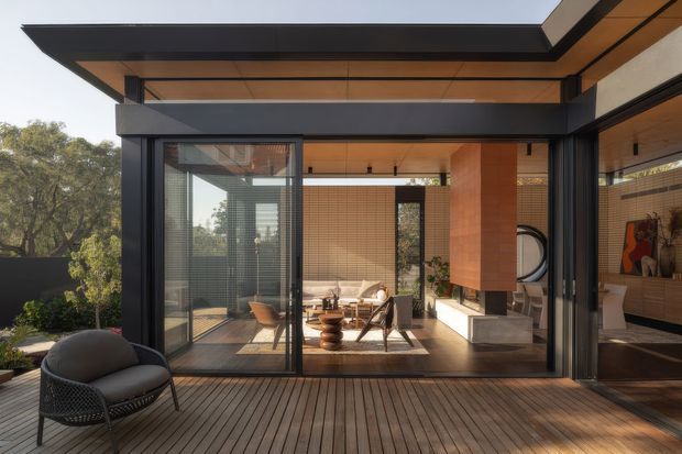 A contemporary pavilion-like living space opening onto a timber deck, framed by expansive black metal glazing and deep charcoal roof beams. Warm timber ceiling lining and terracotta-toned tiled walls surround a furnished lounge with a low coffee table, lounge chairs, and a pale sofa. The image emphasizes indoor-outdoor connection, strong horizontal rooflines, and views toward mature trees.