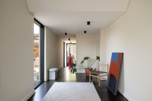 A bright, minimalist residential hallway or entry passage with floor-to-ceiling glazing on one side, white brick wall detailing, dark timber flooring, and a large pale patterned rug. Potted plants, a timber lounge chair, a small side table, and colourful abstract artwork create a restrained contemporary interior. A person is visible near the far doorway, with glimpses of an exterior courtyard beyond.