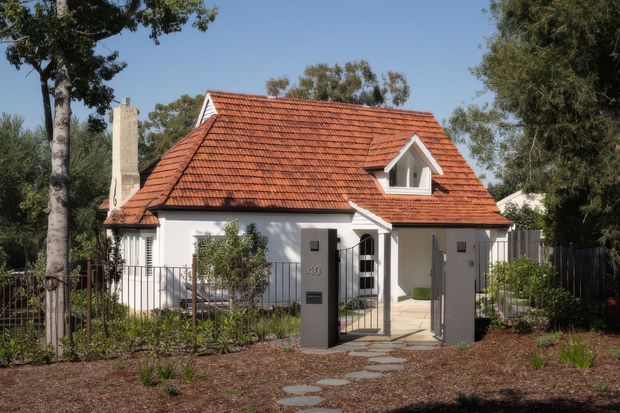 A compact detached house viewed from the front, with white rendered walls, a steep red terracotta tiled gable roof, a small front dormer, and a prominent chimney. A gated paved path leads through a landscaped garden to the recessed entry. Dark metal fencing and gates frame the property, with mature trees around it.