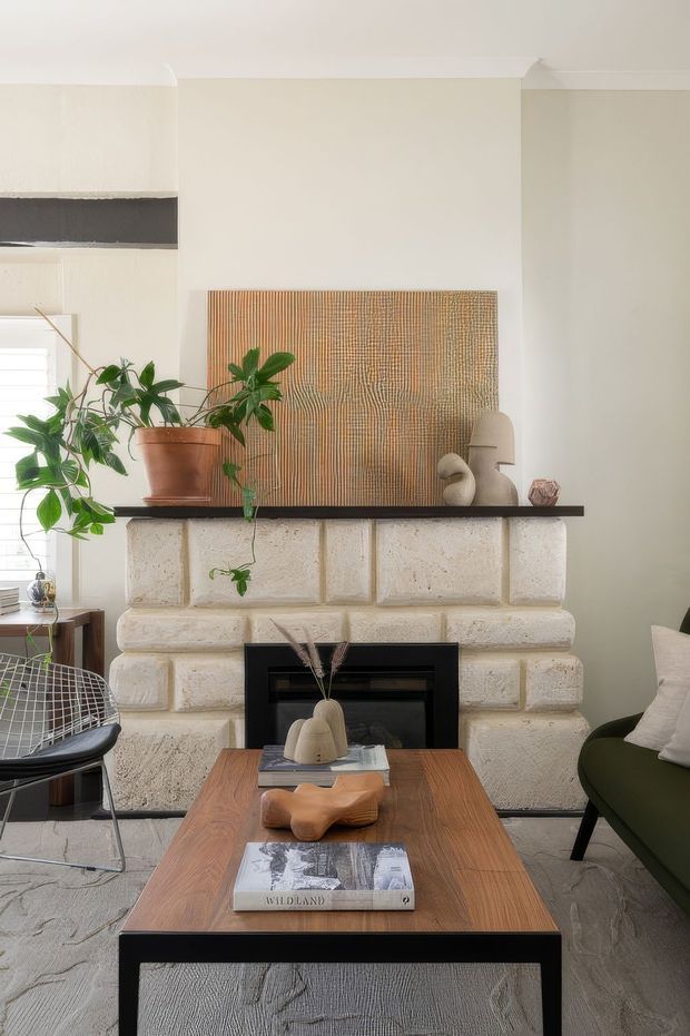 A warm, refined living room centered on a substantial cream masonry fireplace with dark stone or timber mantel and black firebox surround. A large textured abstract artwork rests above the mantel, accompanied by a terracotta potted plant and sculptural decor. The room includes a walnut-toned rectangular coffee table, a dark green upholstered chair, a wire-frame chair, pale walls with traditional cornice detailing, and a light textured rug.
