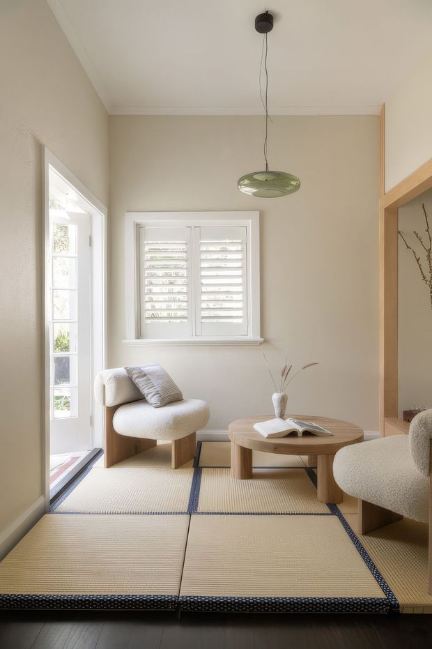 A serene, compact sitting room with Japanese-inspired styling. The room features cream textured walls, white plantation shutters, a glazed door, pale upholstered lounge chairs, a low rounded timber coffee table, and a large tatami-style floor covering with dark edging. A sculptural green pendant light hangs from the ceiling, while a timber-framed opening and built-in shelving appear at right.