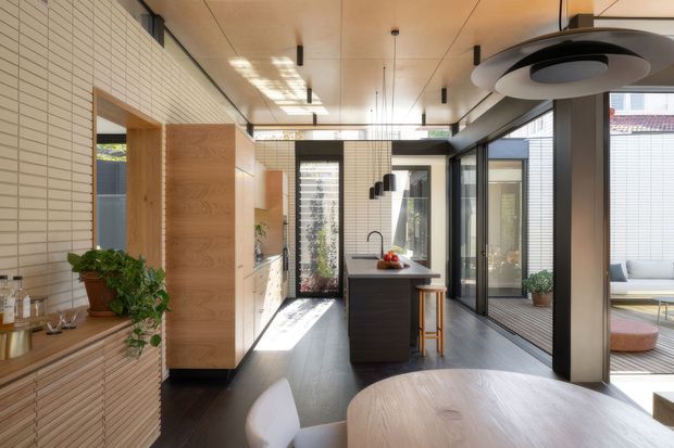 A bright contemporary kitchen and dining area in a narrow, glazed pavilion-like interior. The space features white linear brick wall cladding, warm timber cabinetry and ceiling panels, dark flooring, a compact central island with seating, black-framed floor-to-ceiling glazing, and strong indoor-outdoor connections to a courtyard or terrace. A round timber dining table occupies the foreground, with integrated linear lighting and a sculptural ceiling fixture.