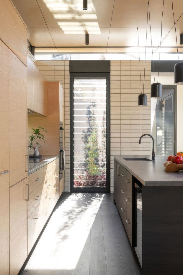 A narrow, contemporary galley kitchen with full-height light timber cabinetry on the left and a dark island with an integrated sink and curved black tap on the right. The space features a pale linear tiled backsplash, dark flooring, exposed ceiling panel joints, black pendant lights, and a tall glazed louvred door or window bringing in strong natural light and views of planting outside.