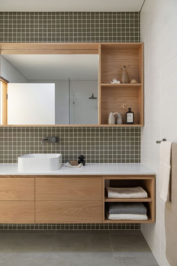 A contemporary bathroom vanity with a large frameless mirrored cabinet, open timber shelving, wall-mounted timber drawers, a white vessel basin, and a dark wall of small square mosaic tiles. White tiled walls, grey floor tiles, a wall-mounted tap, shower visible in the mirror, and neatly folded towels complete the clean, restrained fit-out.