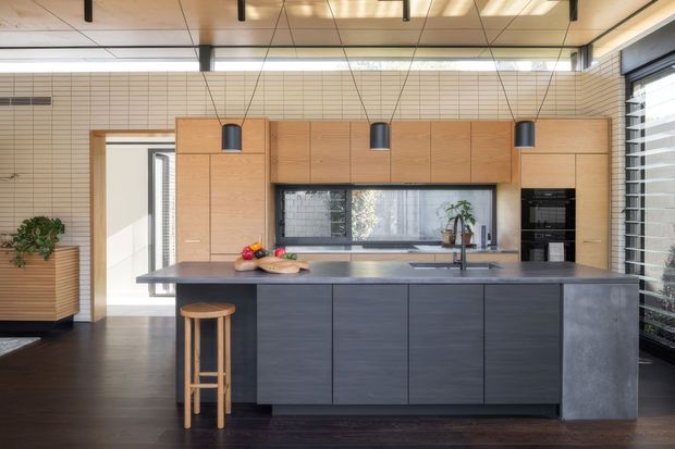 Contemporary kitchen with a large dark island, timber cabinetry, integrated appliances, a wide horizontal window splashback, and a warm timber-lined ceiling. The space combines dark joinery and benchtops with light timber and pale tiled walls, creating a refined, modern interior.