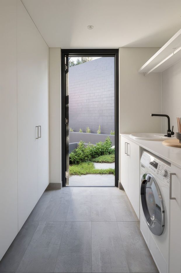 A bright, narrow contemporary laundry with full-height white cabinetry on the left and built-in white cabinetry on the right, incorporating a front-loading washing machine, white utility sink and matte black tapware. Large grey floor tiles continue toward an open black-framed glass door leading to a small planted courtyard. White walls and ceiling, streamlined joinery and open overhead shelving create a clean, minimalist look.