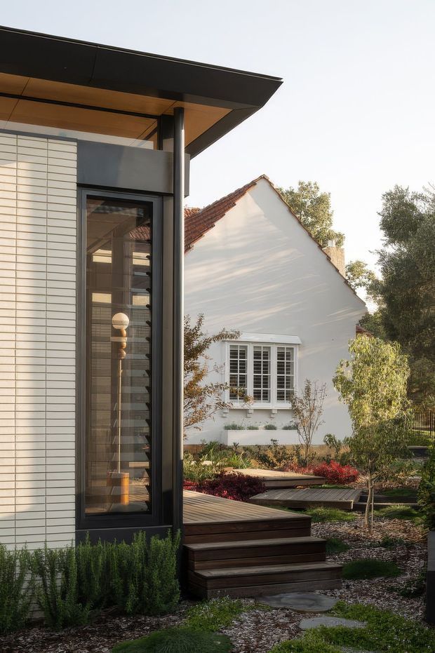Exterior architectural photograph showing a contemporary extension beside an older white gabled house. The foreground structure has crisp white horizontal cladding, dark metal-framed glazing, a broad projecting flat roof with warm timber soffit, and a raised timber deck with steps leading into a landscaped garden. The background dwelling features a traditional tiled gable roof and multi-pane window, highlighting the contrast between new and existing architecture.