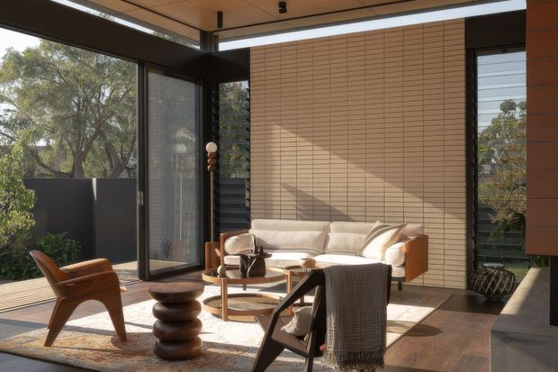 A sunlit contemporary living room or sunroom with floor-to-ceiling glazing opening to a leafy outdoor setting. The space features warm timber ceiling and wall paneling, dark metal-framed windows and doors, a cream sofa, sculptural wooden coffee table, leather lounge chair, woven throw, and a neutral patterned rug. Strong natural light creates a calm, warm, refined atmosphere.