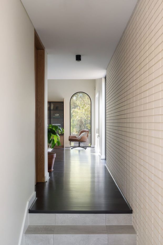 A bright, narrow residential hallway viewed from a tiled stair landing. The corridor has dark timber flooring, a long cream brick-tile feature wall on the right, white plaster walls and ceiling, and timber door surrounds. At the far end, an arched window brings in natural light beside a contemporary lounge chair and indoor plant, with glazed cabinetry visible in an adjoining room.