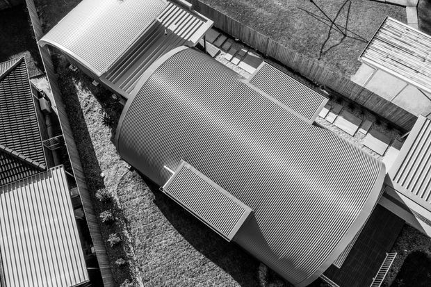 A black-and-white aerial view of a contemporary building complex, dominated by a large curved corrugated-metal roof with several adjoining smaller roof forms, covered walkways, and landscaped edges. The image focuses on the roof geometry and overall site composition rather than an interior space.