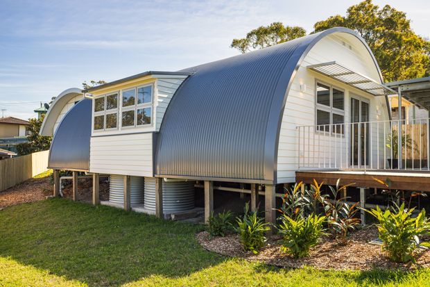 Exterior view of a distinctive elevated single-storey dwelling with a curved barrel-vault roof and corrugated metal cladding. The facade combines dark grey curved roofing with white horizontal weatherboard-style wall panels, white-framed windows, and a raised timber deck with pale metal balustrades. Garden planting and lawn surround the structure, with storage or service elements visible beneath the elevated floor.