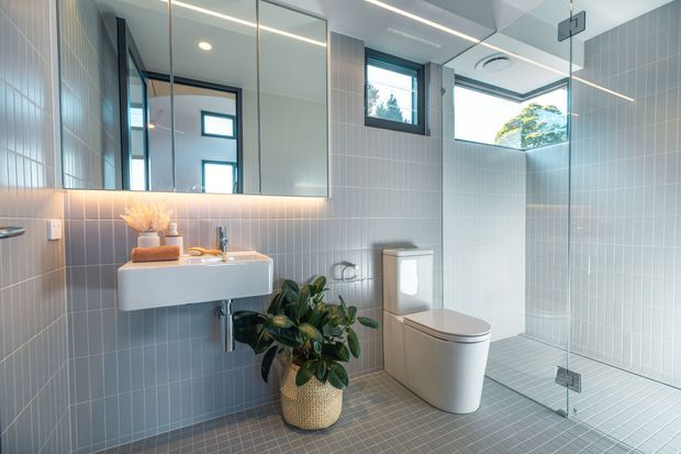 A bright contemporary bathroom with pale blue-grey vertical wall tiles, matching small-format floor tiles, a floating white vanity with integrated basin, wall-mounted tapware, large mirrored cabinetry, and a white toilet. A frameless glass shower enclosure occupies the right side, with a high window providing natural light. Recessed and linear lighting, black-framed windows, and a potted plant add modern detailing.