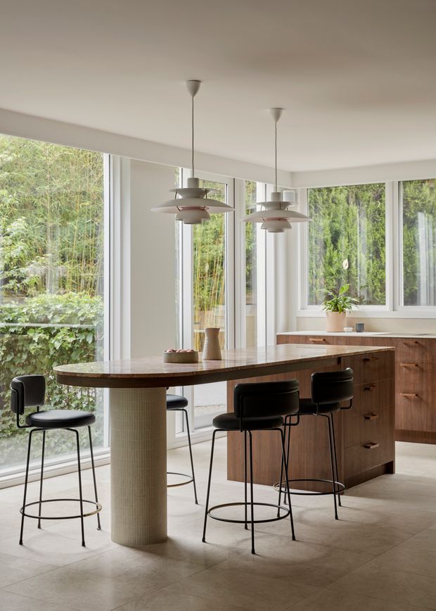 A bright, contemporary kitchen with a large curved island and rounded tiled support column, dark timber cabinetry, pale stone or tile flooring, and black bar stools. Two sculptural pendant lights hang above the island. Extensive floor-to-ceiling glazing opens the room to lush greenery, creating an airy indoor-outdoor atmosphere.