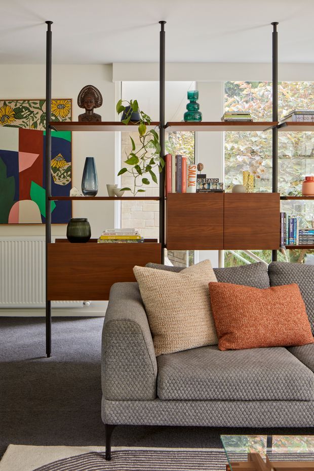 A warmly styled living room with a grey textured sofa in the foreground, layered beige and rust cushions, dark carpet, and a glass coffee table. A freestanding open shelving and cabinetry unit in dark timber with slim black metal supports divides the room, displaying books, ceramics, plants, and sculpture. Abstract and botanical artwork, a radiator, large windows, and leafy outdoor views are visible in the background.