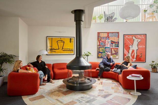 Bright contemporary living room with a suspended black cylindrical fireplace at its centre, surrounded by a curved red sectional sofa and matching lounge chairs. The room features a pale patterned tiled or stone floor, white walls, colourful framed artwork, indoor plants, a sculptural pendant light, and an elevated mezzanine or stair balustrade visible above.